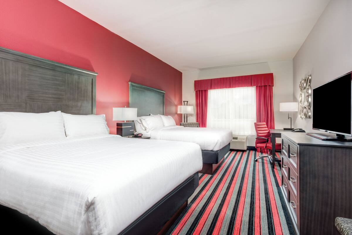 A hotel room with two beds, a desk with a TV, a chair, and a red-striped carpet.
