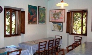 A dining area with a table and chairs, framed artwork on the walls, and windows with open shutters. - Agriturismo Su Tiresi (Dorgali)