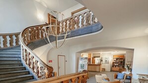 Una amplia escalera con barandillas ornamentadas que conduce a un segundo piso, una zona de recepción con mostrador y asientos, y una espaciosa sala de estar con chimenea.