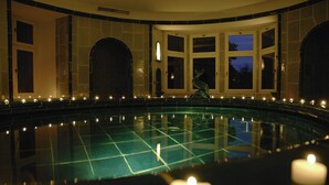 Una amplia piscina interior con suelo de baldosas, rodeada de paredes con ventanales de arco y velas.