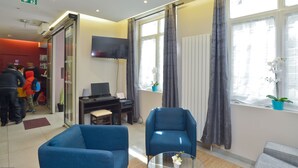 Un vestíbulo de hotel con mostrador de recepción, un televisor de pantalla plana, dos sillones azules y ventanas grandes con cortinas.