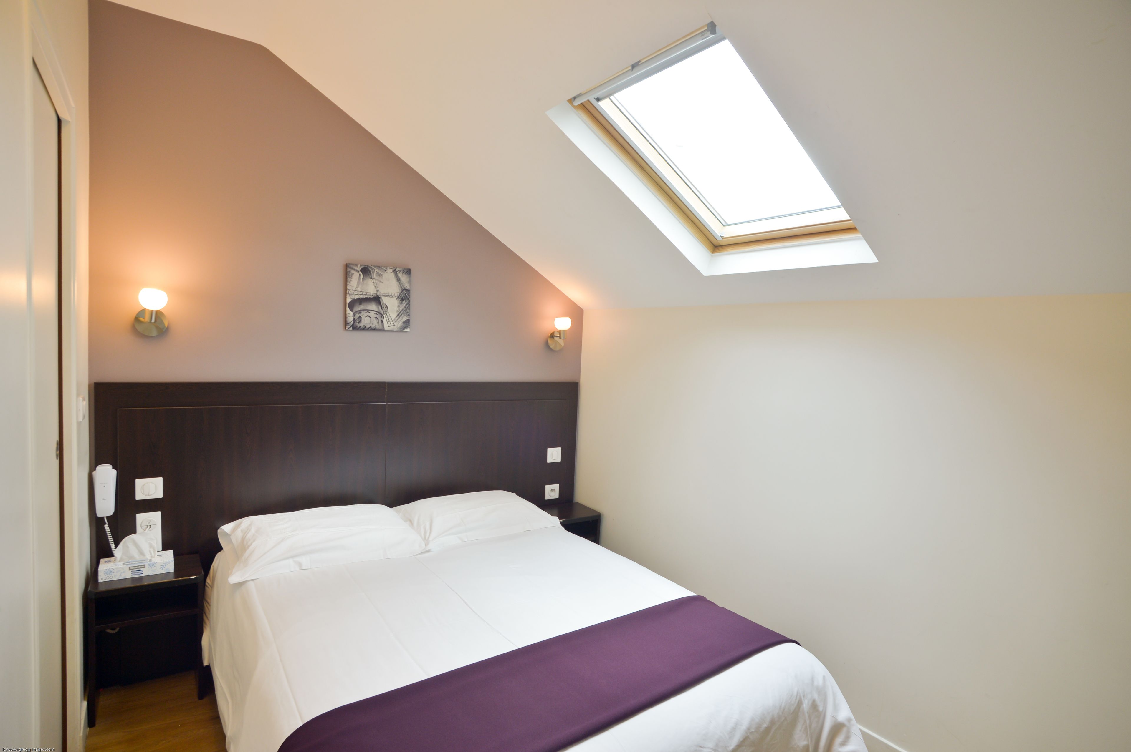 Ein Schlafzimmer mit Bett, Nachttisch, Nachttischlampe, Wandleuchte und Dachfenster.
