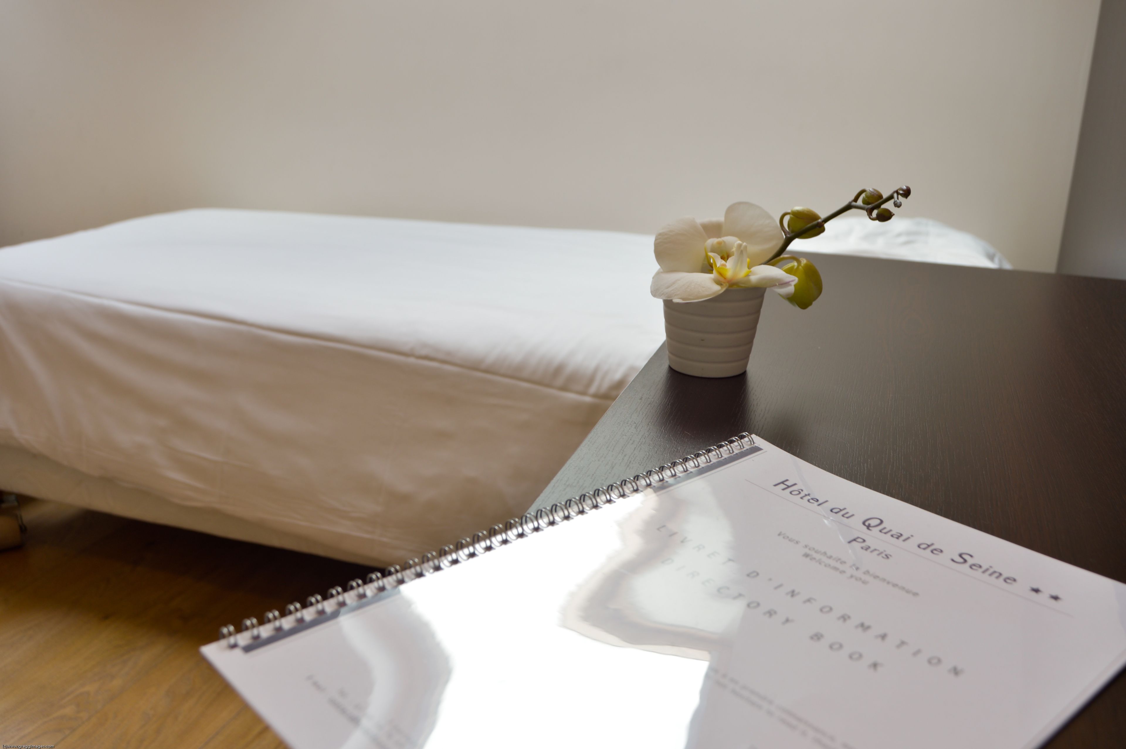 Ein Hotelzimmer mit einem ordentlich bezogenen Bett, einem kleinen Tisch mit einer weißen Orchidee und einer Broschüre darauf.