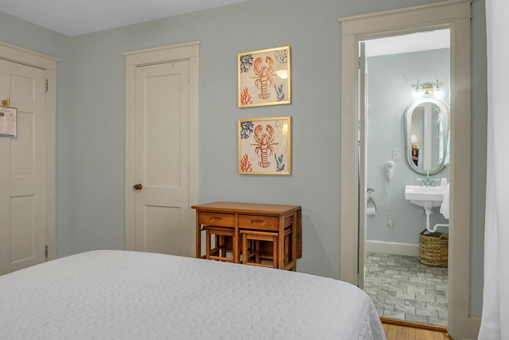 Une chambre à coucher avec un lit, une table de chevet en bois, deux tableaux encadrés accrochés au mur et une salle de bain visible à travers une porte ouverte.