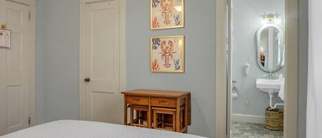 Une chambre à coucher avec un lit, une table de chevet en bois, deux tableaux encadrés accrochés au mur et une salle de bain visible à travers une porte ouverte.