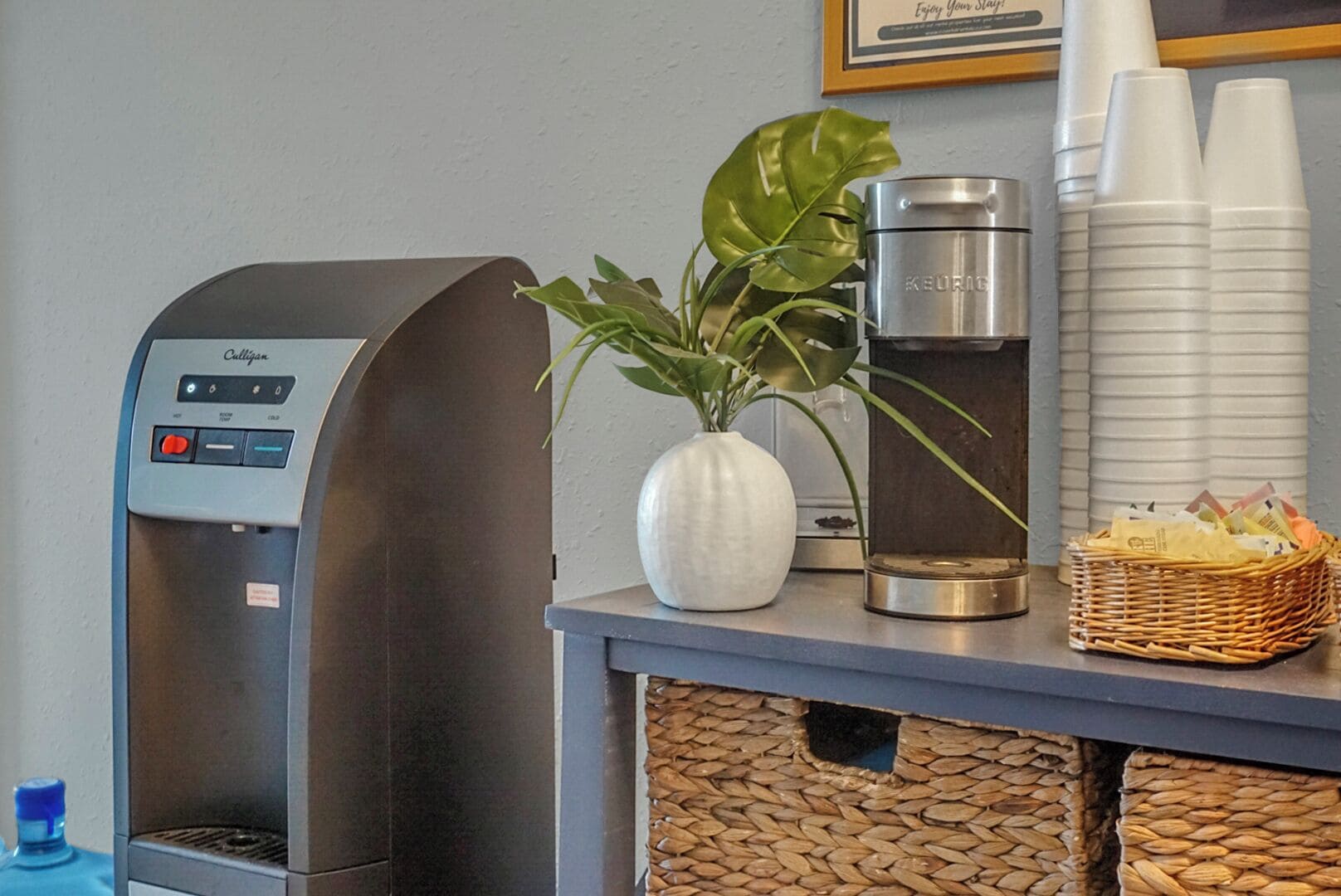 Una cafetera, un dispensador de agua, una planta y una cesta con aperitivos sobre una encimera.