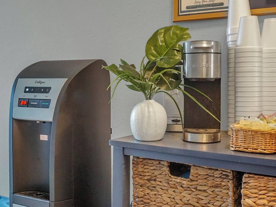 Een koffieapparaat, een waterdispenser, een plant en een mand met snacks op een aanrecht.