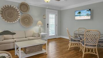 A living room with a sofa, coffee table, and wicker chairs. There is a ceiling fan, a lamp, and a painting of a beach scene on the wall.