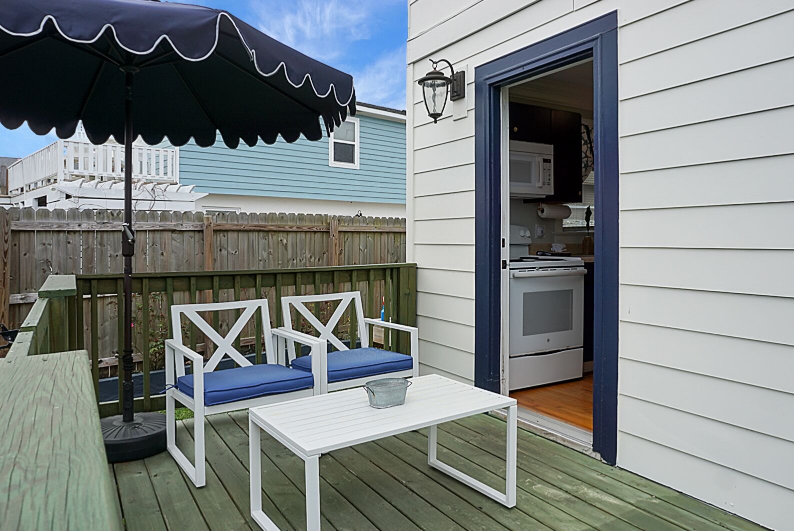 Eine Terrasse mit einem weißen Tisch, zwei blauen Sitzgelegenheiten mit Kissen, einem schwarzen Sonnenschirm und einem Holzzaun im Hintergrund.
