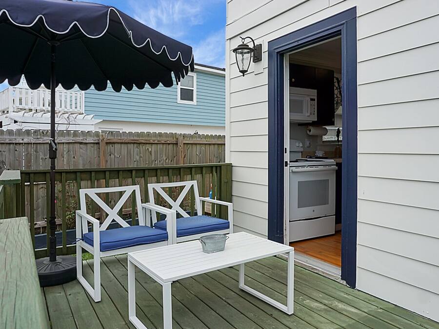 Eine Terrasse mit einem weißen Tisch, zwei blauen Sitzgelegenheiten mit Kissen, einem schwarzen Sonnenschirm und einem Holzzaun im Hintergrund.