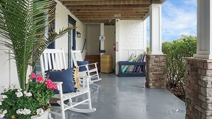 A covered porch with white rocking chairs, a small table, and a stone wall. - Carolina Beach Inn (Carolina Beach)