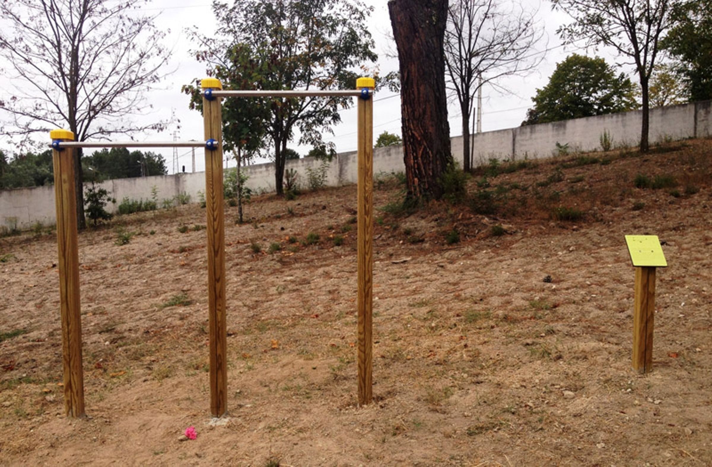 A playground with a wooden structure, including a horizontal bar and vertical poles, set in a natural, outdoor environment with trees and a dirt ground.