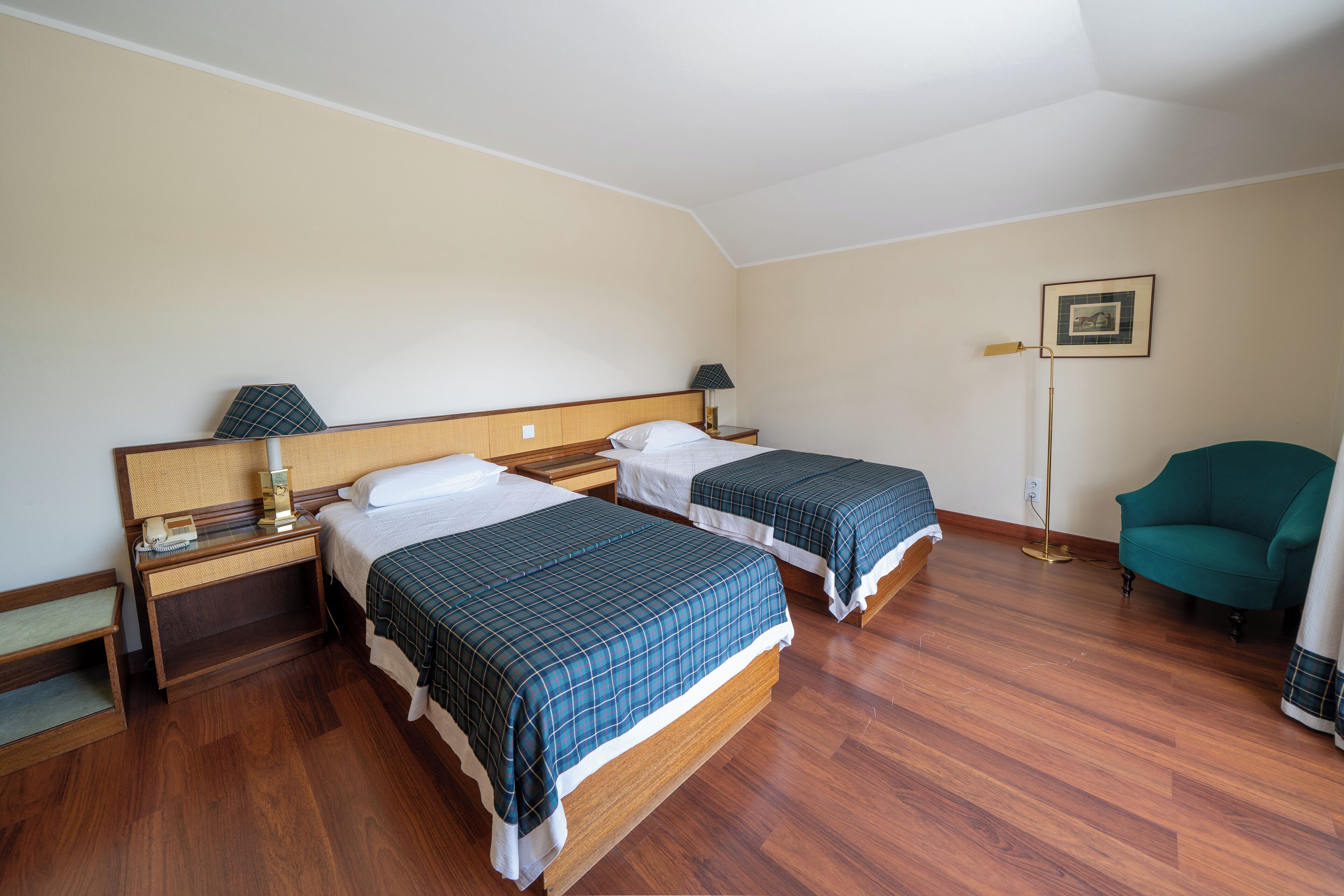A hotel room with two beds, wooden flooring, a green armchair, and a framed picture on the wall.