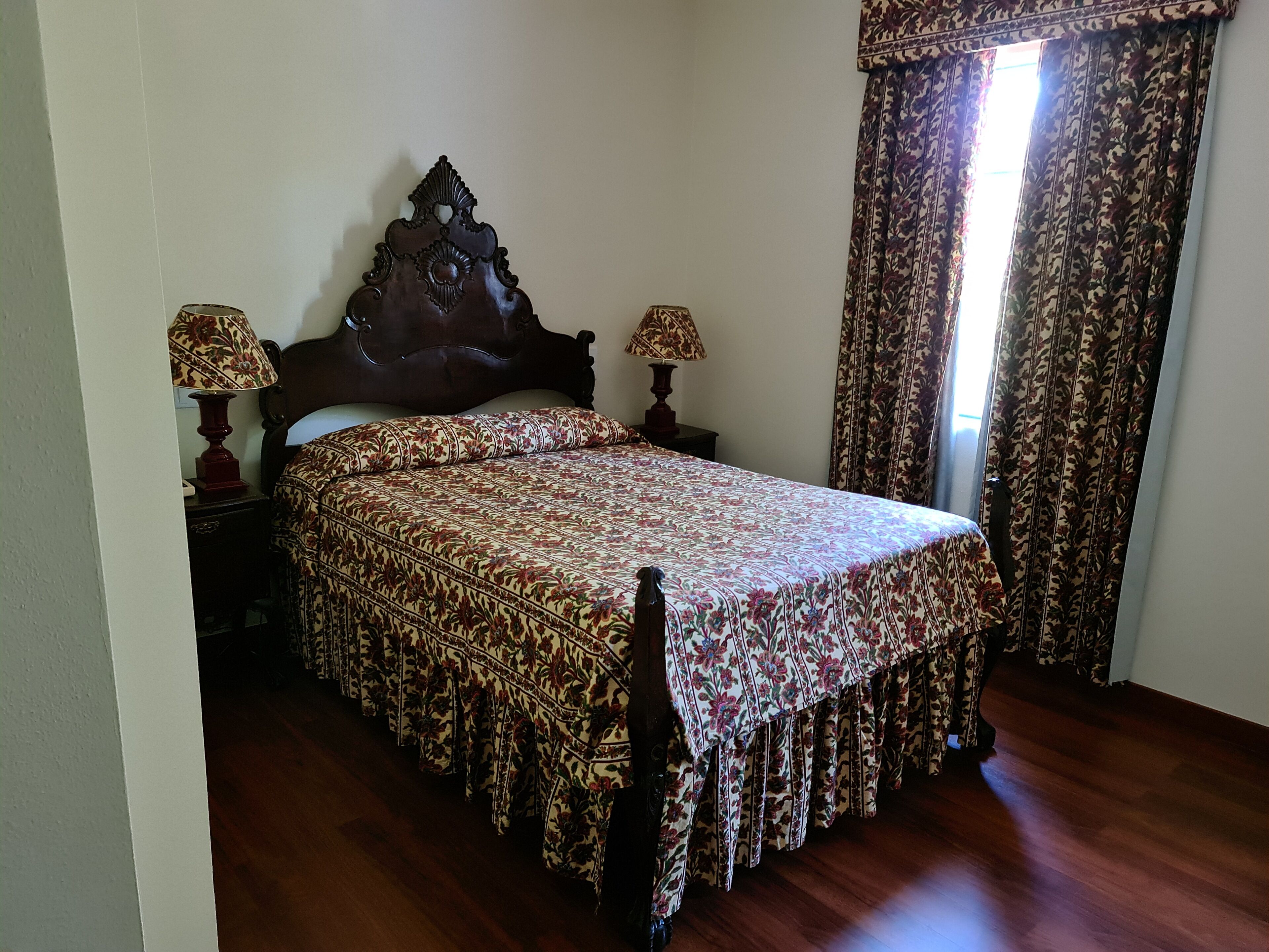 Une chambre à coucher avec un lit en bois, une couvre-lit à motifs floraux, deux lampes de chevet et une fenêtre avec des rideaux.