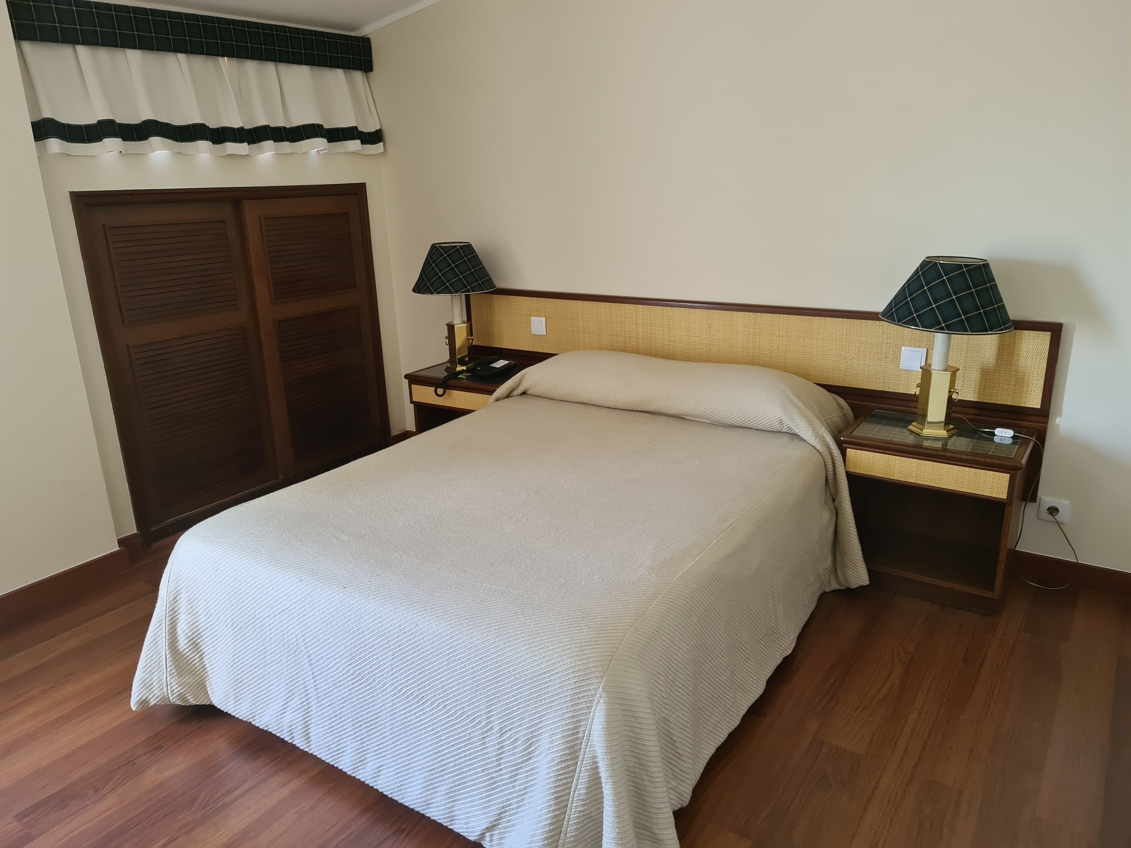 A neatly made bed with a beige bedspread, two bedside tables with lamps, a wooden wardrobe, and a window with a green and white valance.