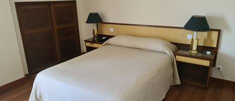 A neatly made bed with a beige bedspread, two bedside tables with lamps, a wooden wardrobe, and a window with a green and white valance.
