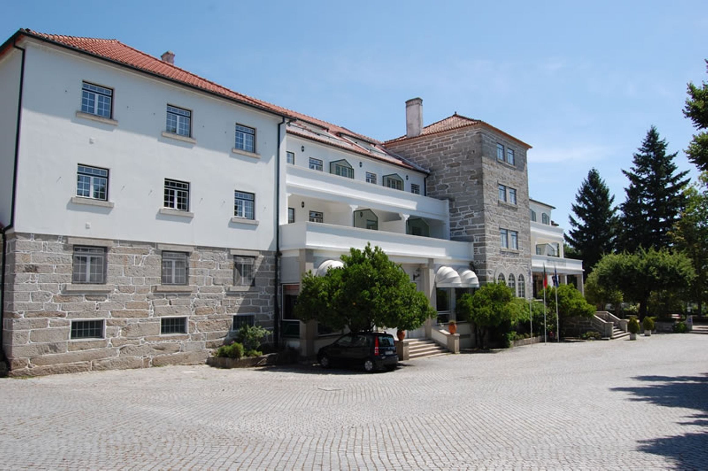 A large building with a stone facade and a white upper section. There is a car parked in front, and a tree is visible to the right.
