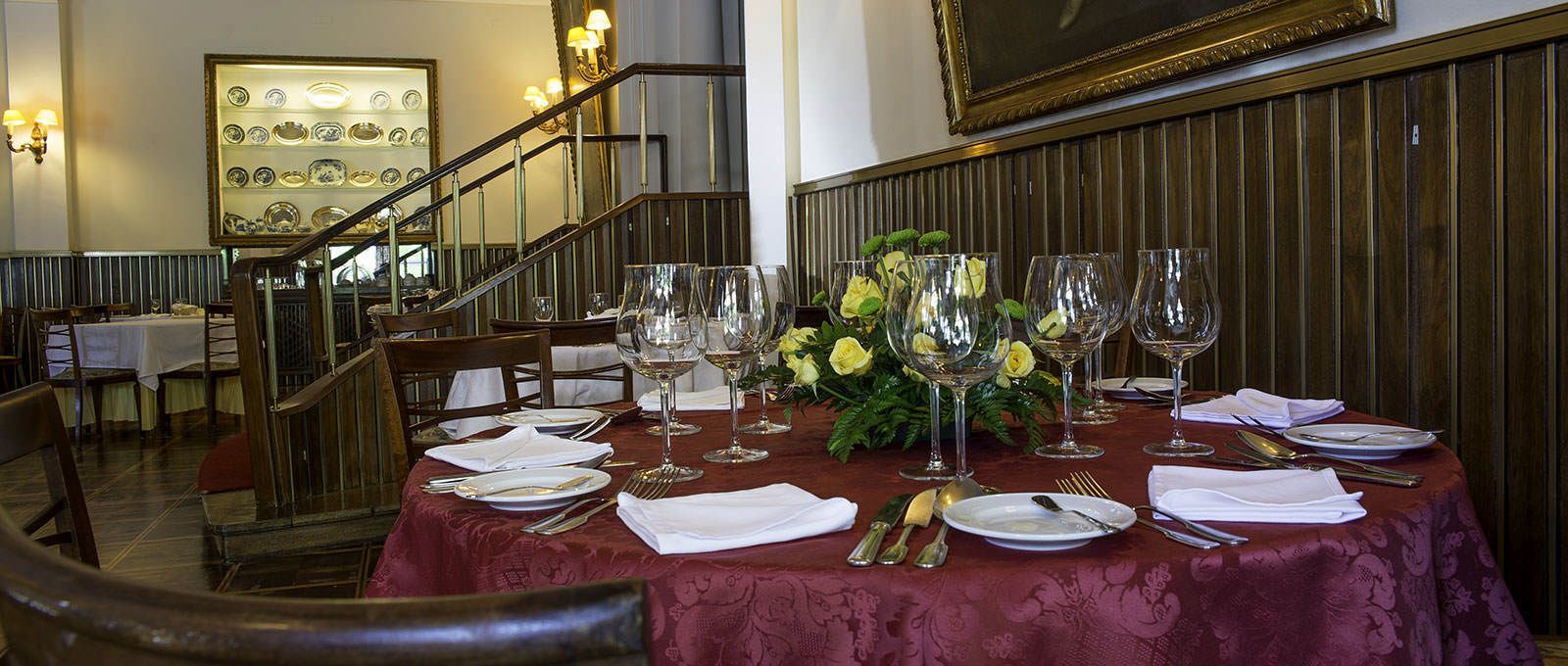 Mesa de jantar posta com toalhas brancas, copos de vinho e talheres, rodeada por cadeiras em restaurante com decoração em madeira e escadaria.