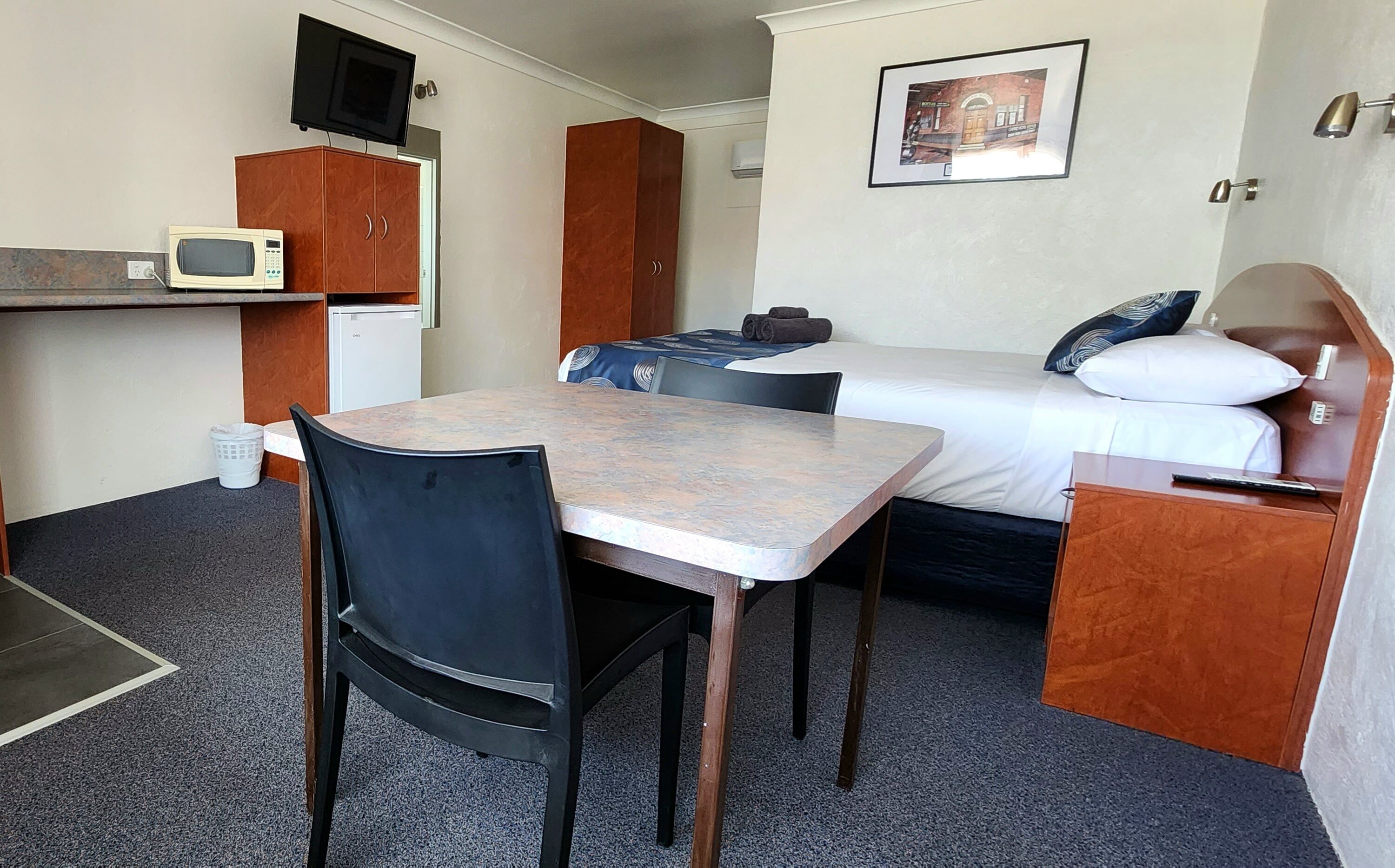 Habitación de hotel pequeña con una cama individual, una mesita con una silla, un televisor montado en la pared, un microondas y un armario pequeño.