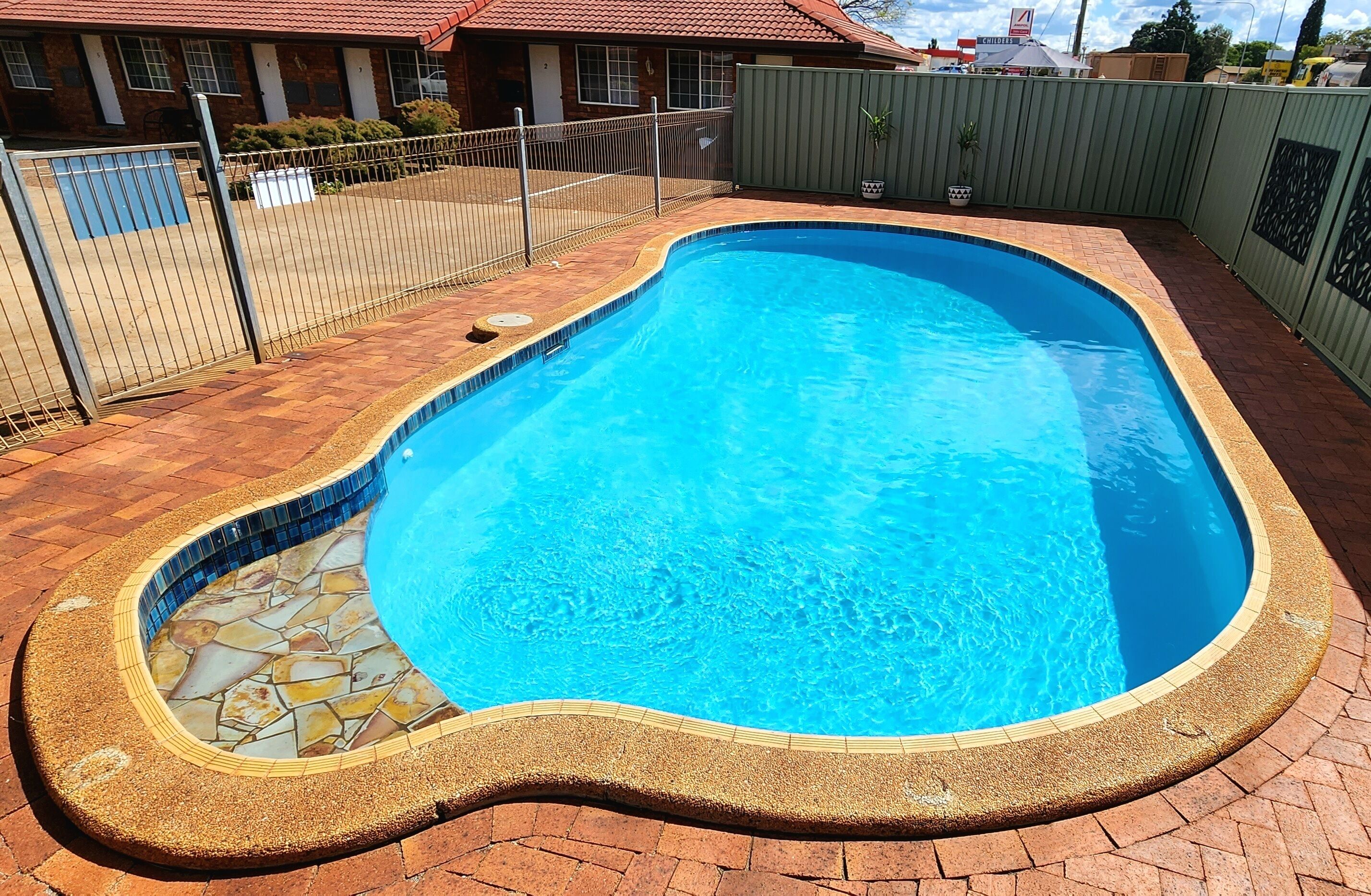 A swimming pool with a tiled edge and a pebble border, surrounded by a brick patio and a metal fence.