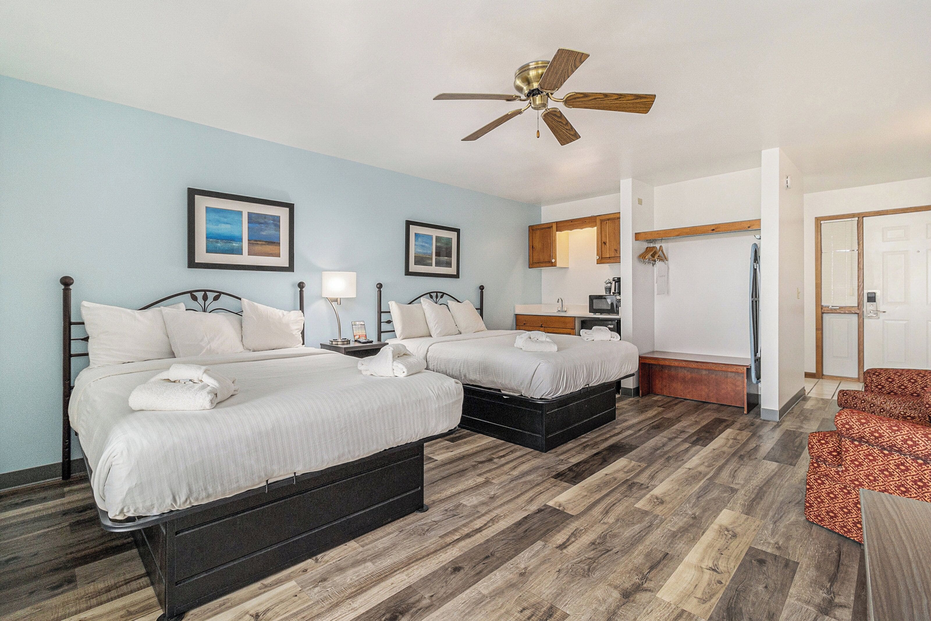 A hotel room with two beds, a ceiling fan, wooden flooring, a small kitchenette, and a bathroom with a shower.