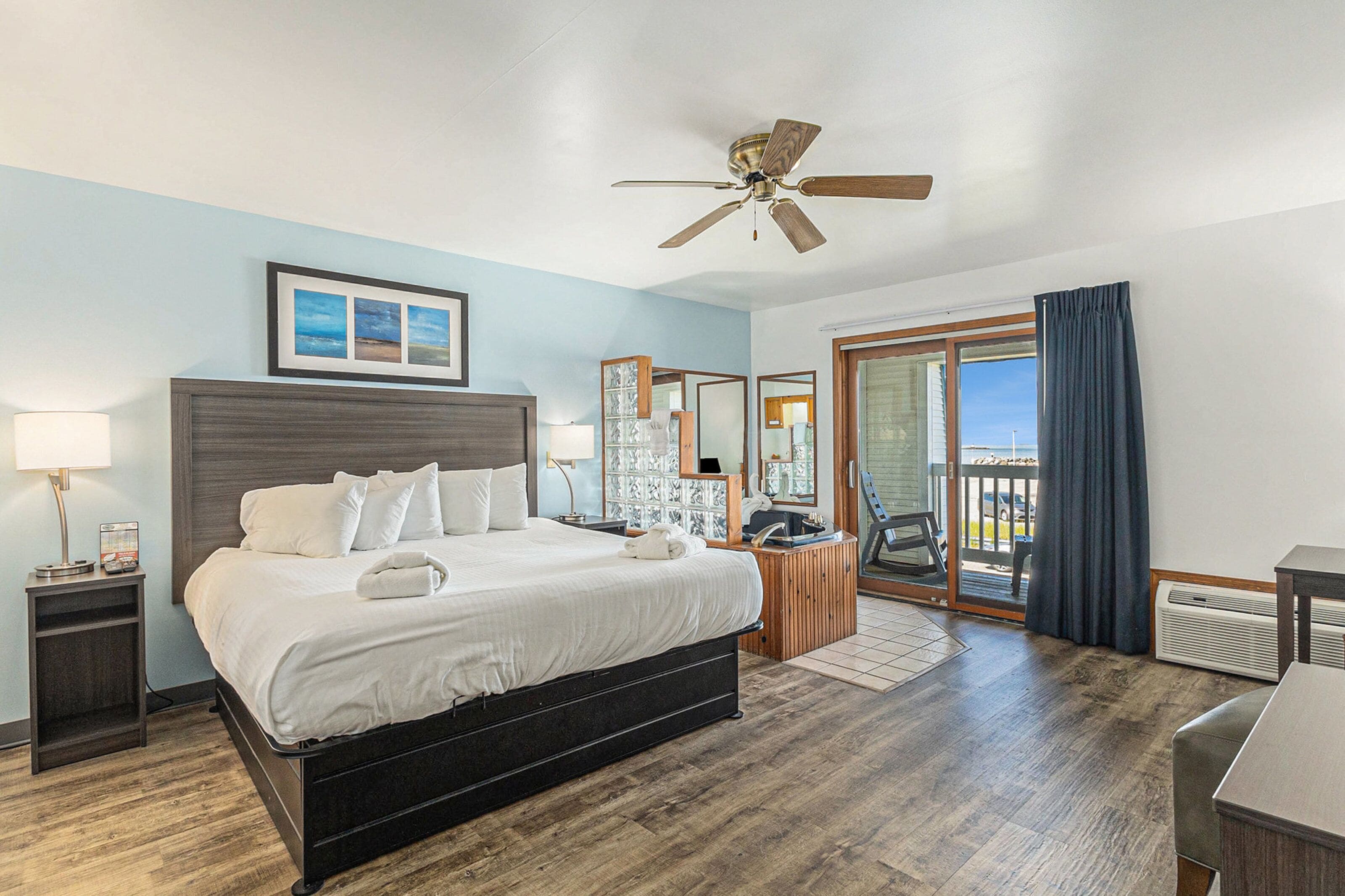 A hotel room with a bed, a ceiling fan, a large window with a view, and a door leading to a balcony.