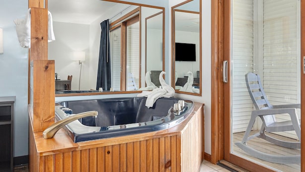 A bathroom with a wooden bathtub, a large mirror, and a glass door leading to a balcony with a chair.