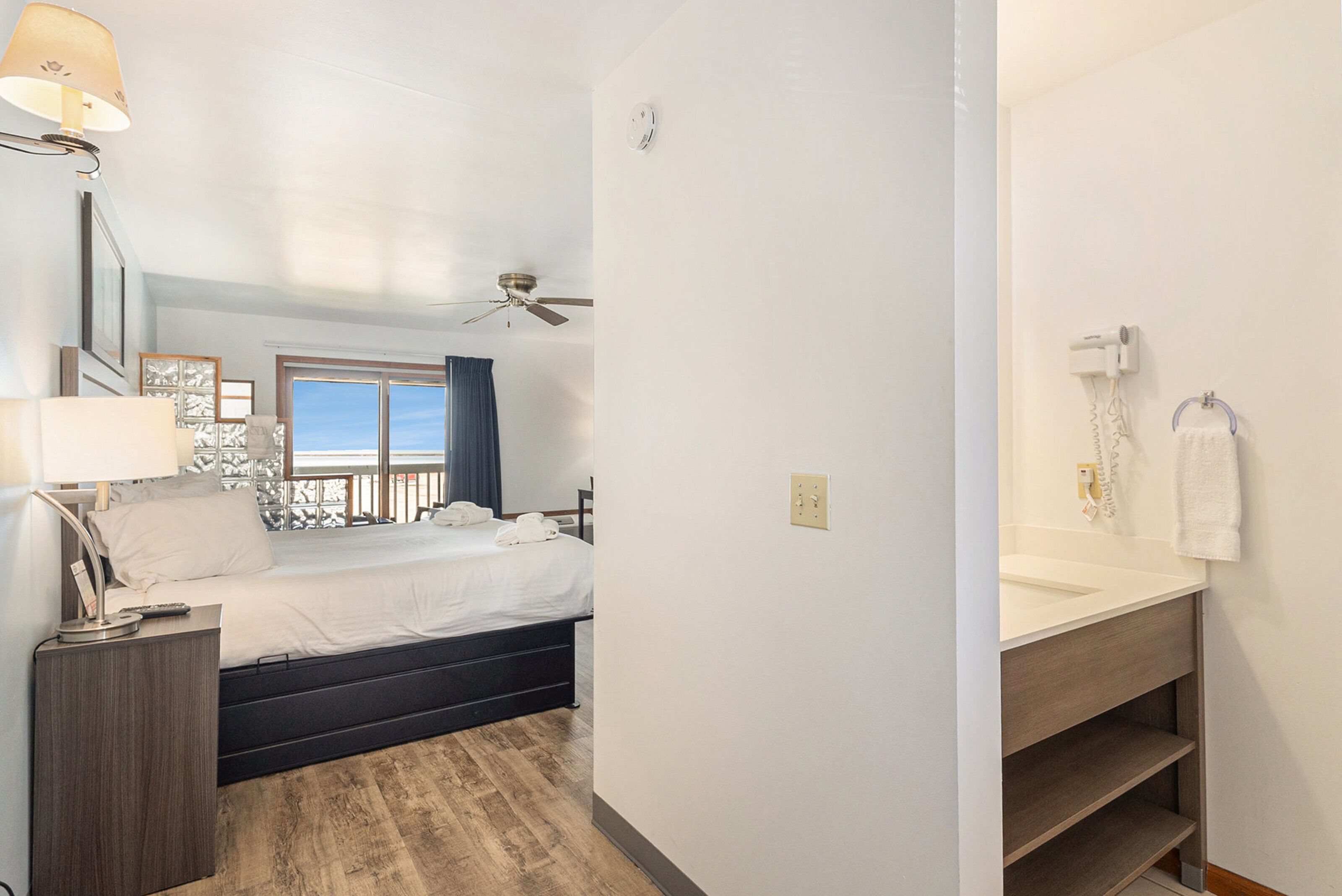 A hotel room with a bed, bedside table, lamp, ceiling fan, window with curtains, and a bathroom with a sink and towel rack.