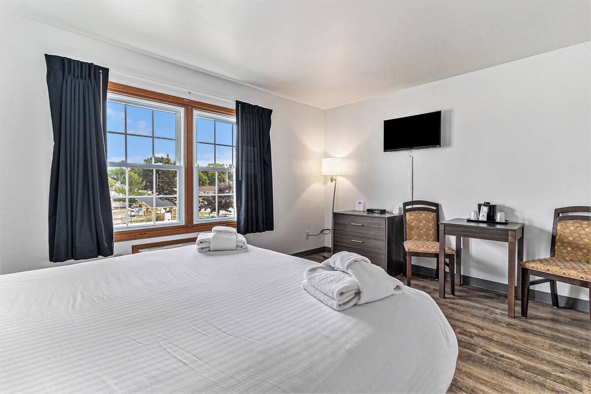Quarto de hotel com uma cama grande, uma escrivaninha com cadeira, uma televisão fixada na parede e uma janela com cortinas.