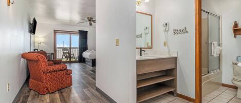 A hotel room with a wooden floor, a red patterned sofa, a bathroom with a shower, and a balcony with a view.