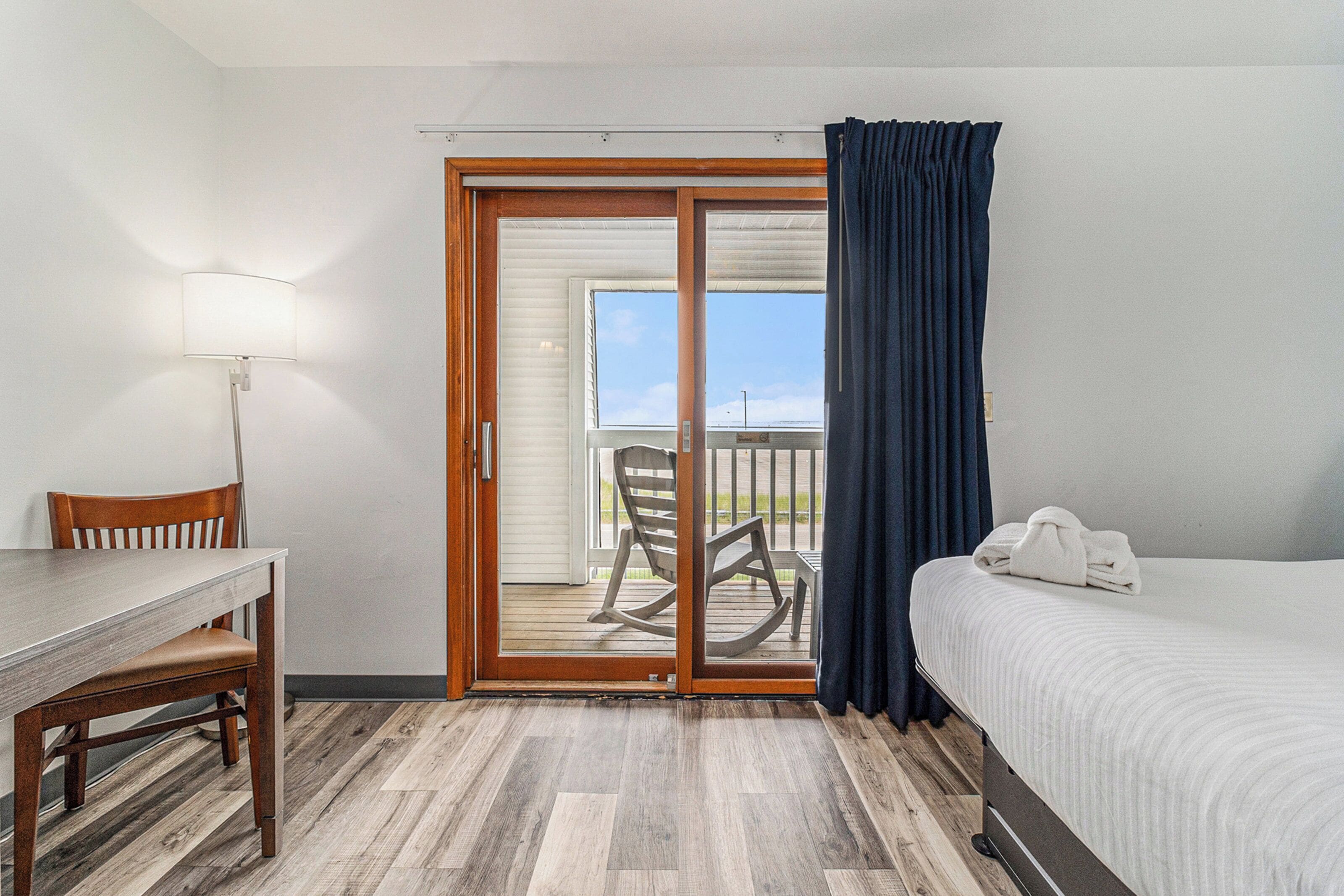 A hotel room with a bed, a desk, and a chair. There is a sliding glass door leading to an outdoor area with a chair and a table.