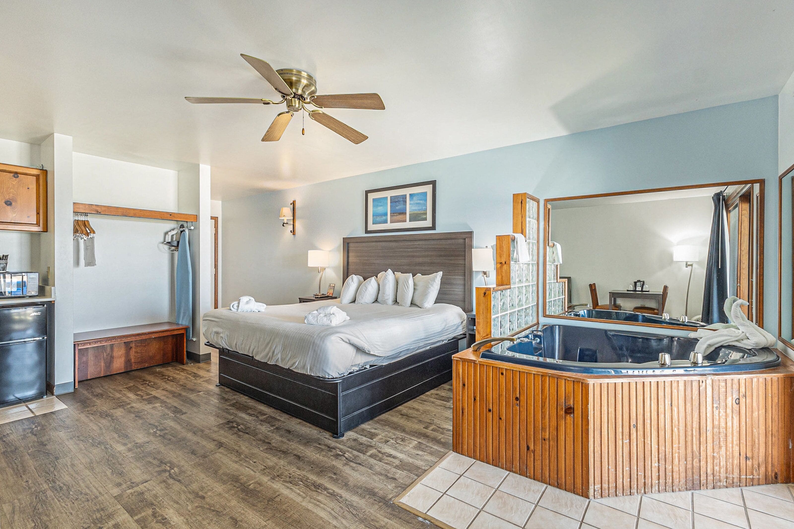 A hotel room with a bed, a wooden bench, a ceiling fan, a large mirror, and a hot tub.
