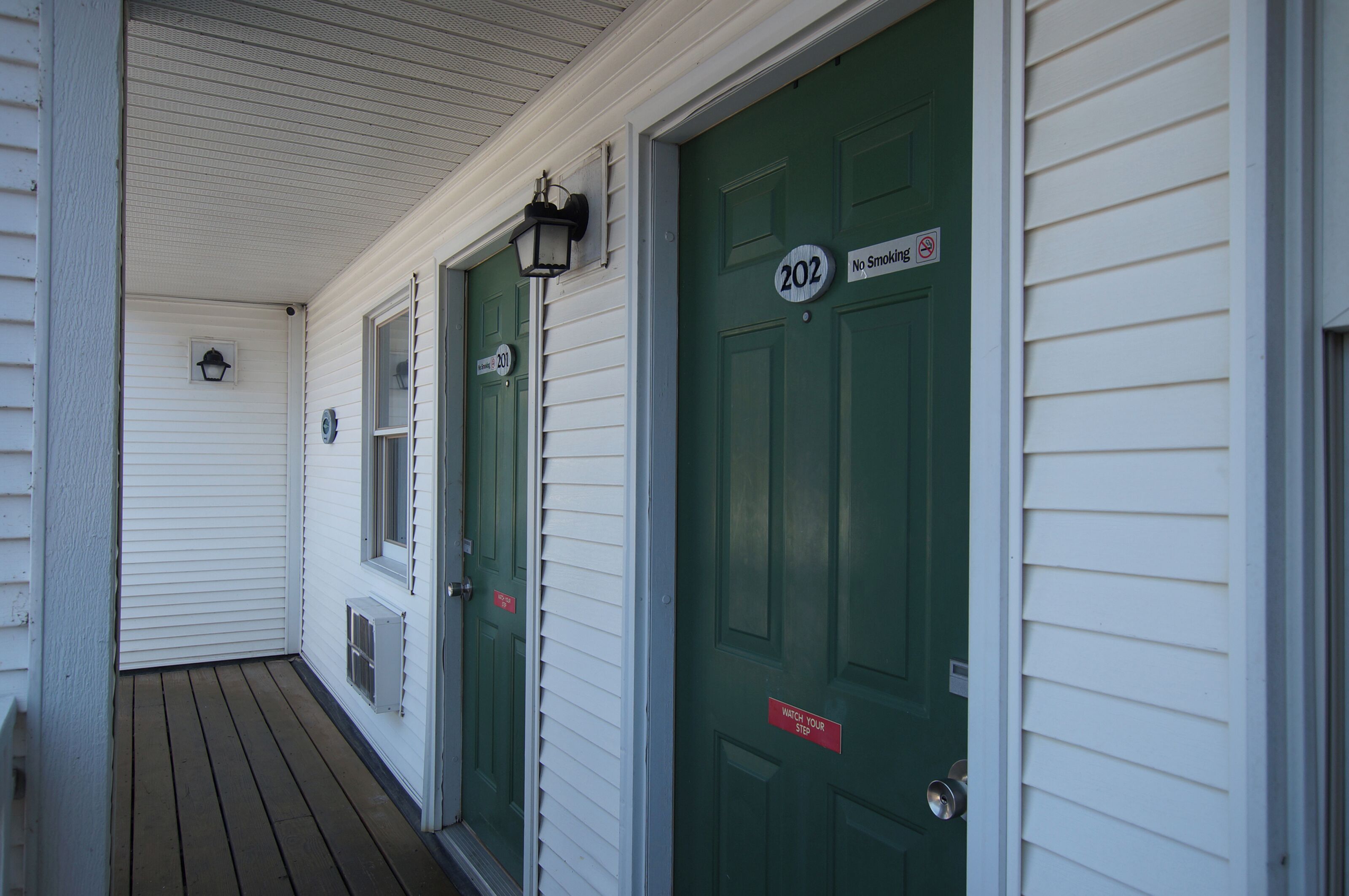 A building with green doors labeled 202 and 203, a porch with a wooden floor, and a small window with a light fixture.