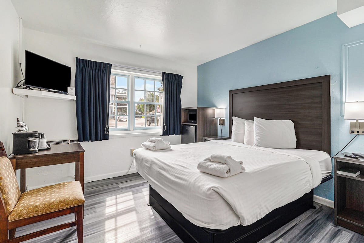 Quarto de hotel com uma cama grande, televisão fixada na parede, escrivaninha com candeeiro e vista para o exterior pela janela.