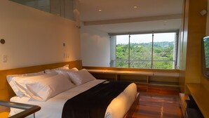 Ein modernes Hotelzimmer mit einem großen Bett, einer Bank am Fußende und Holzboden, mit einem großen Fenster und Blick auf Grünflächen.