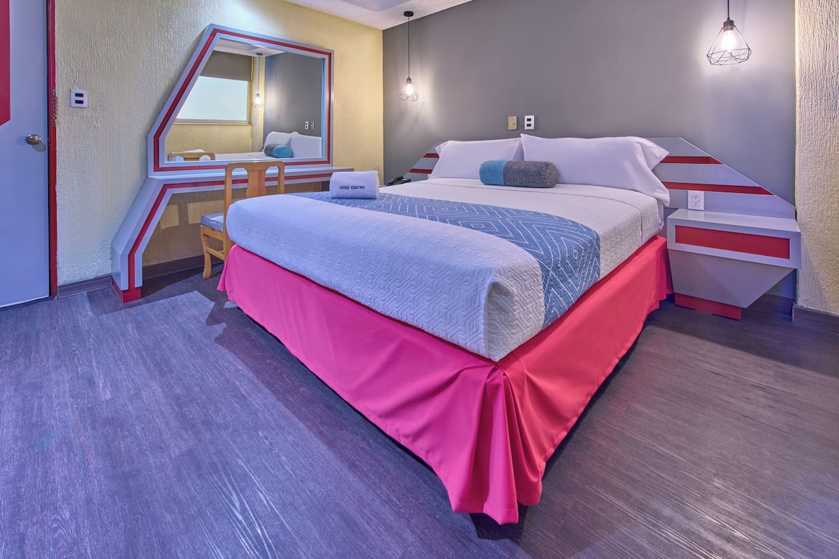 Hotellihuone, jossa on sänky, työpöytä, peili ja punavalkoraidallinen yöpöytä.