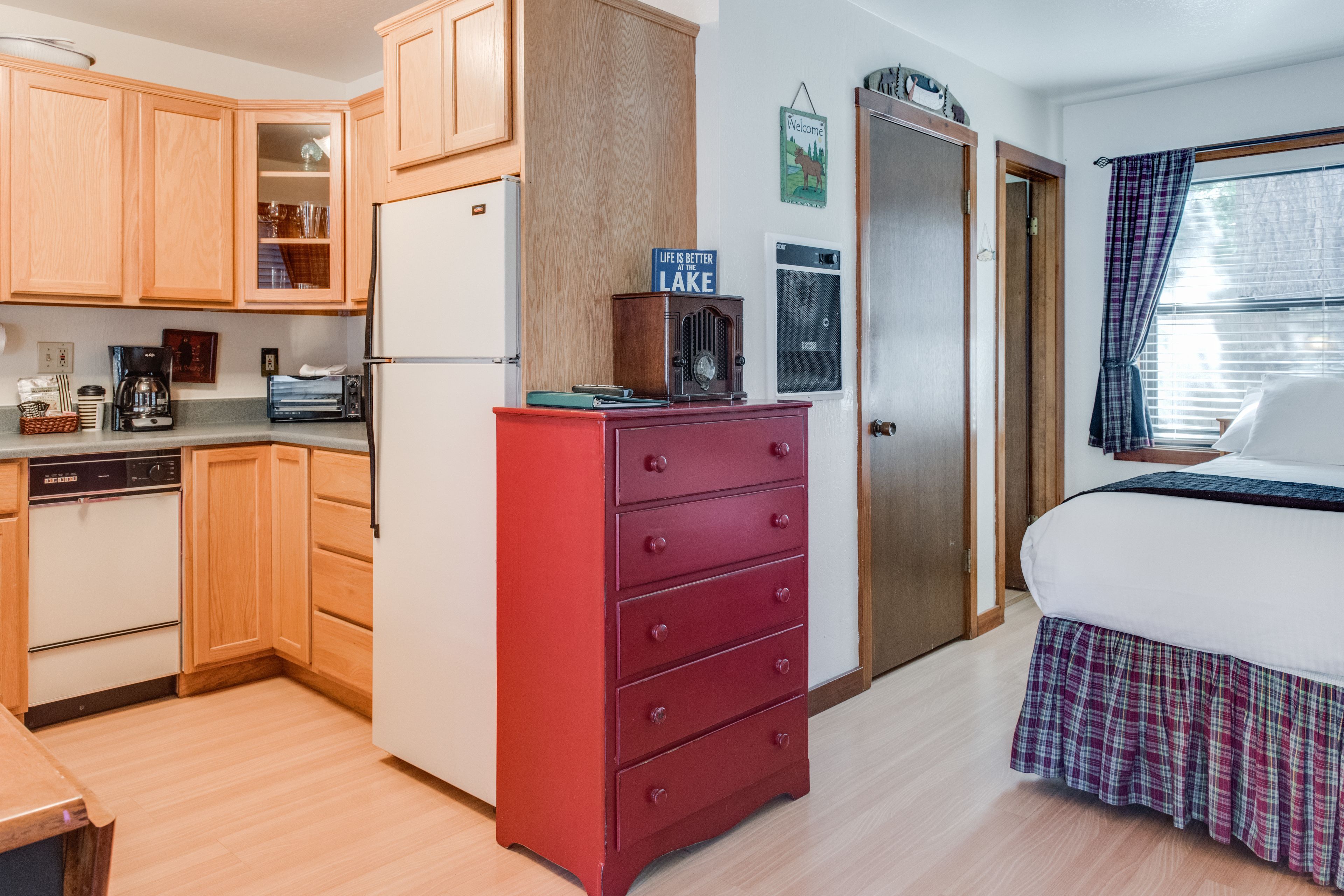 A compact kitchen with wooden cabinets, a red dresser, and a bed with a plaid skirt.