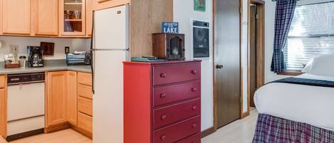 A compact kitchen with wooden cabinets, a red dresser, and a bed with a plaid skirt.
