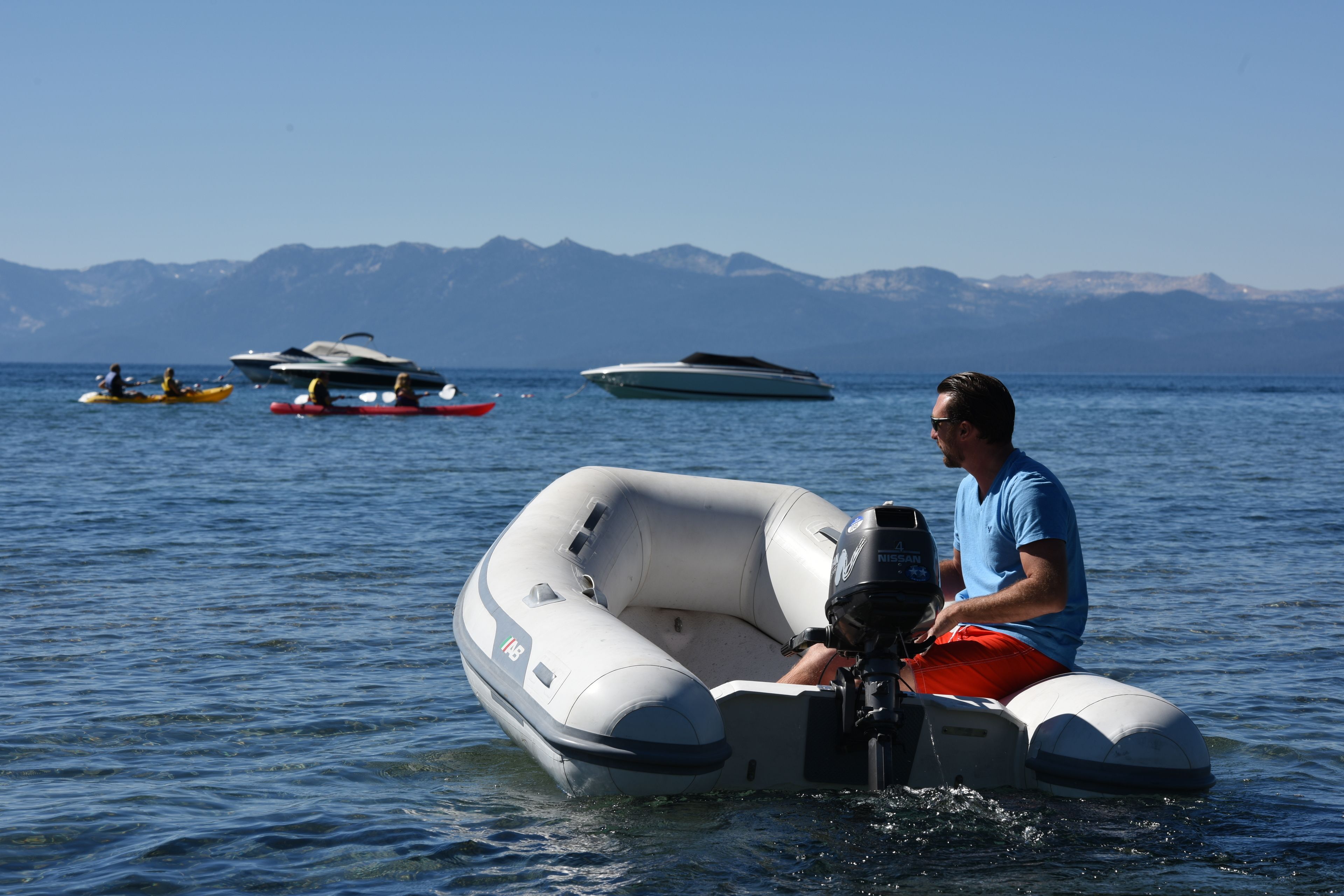 A man in a blue shirt and red shorts on a motorized inflatable boat on calm water.