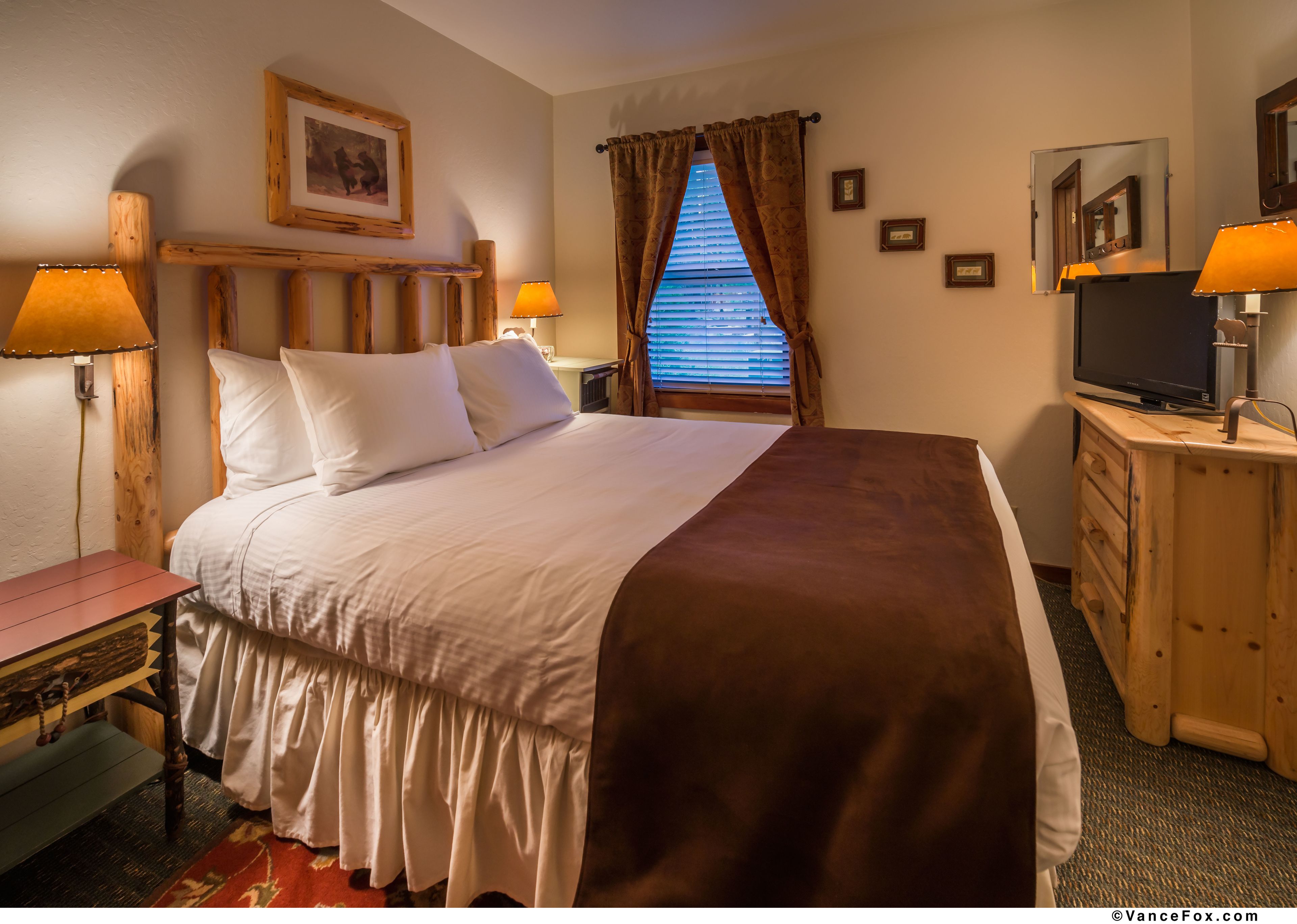 A neatly made bed with white and brown bedding, a wooden headboard, a bedside table with a lamp, a dresser with a TV, and a window with curtains.