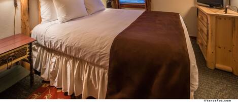 A neatly made bed with white and brown bedding, a wooden headboard, a bedside table with a lamp, a dresser with a TV, and a window with curtains.