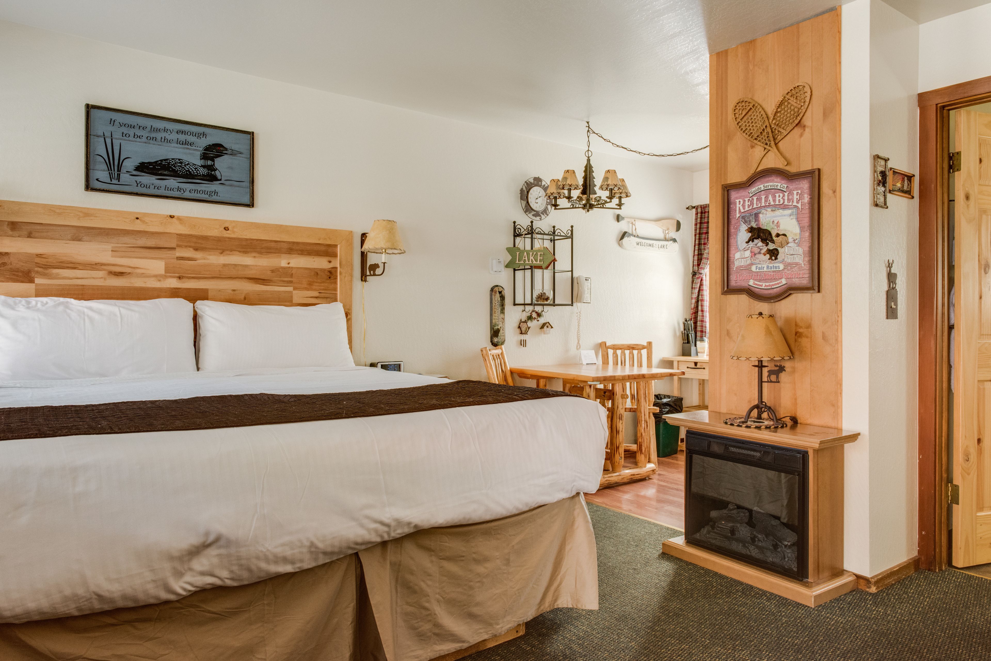 Un dormitorio con cabecera de madera, cama con sábanas blancas, una mesita de madera con sillas, una lámpara de pared y una chimenea.