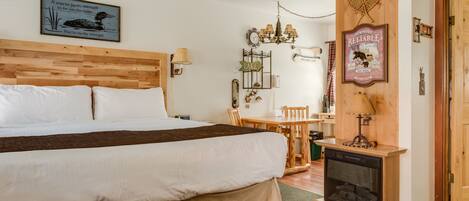 Un dormitorio con cabecera de madera, cama con sábanas blancas, una mesita de madera con sillas, una lámpara de pared y una chimenea.