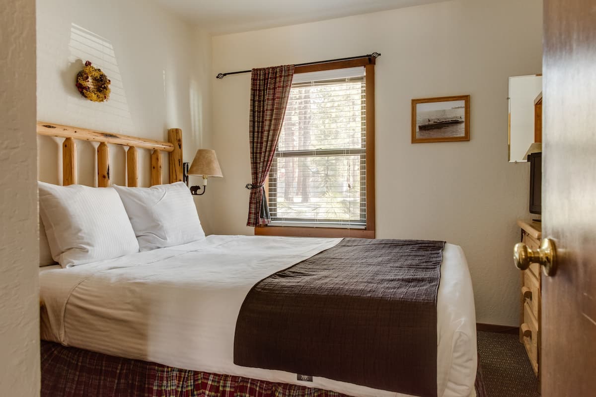 A neatly made bed with a headboard, a window with curtains, a wall-mounted lamp, and a framed picture.