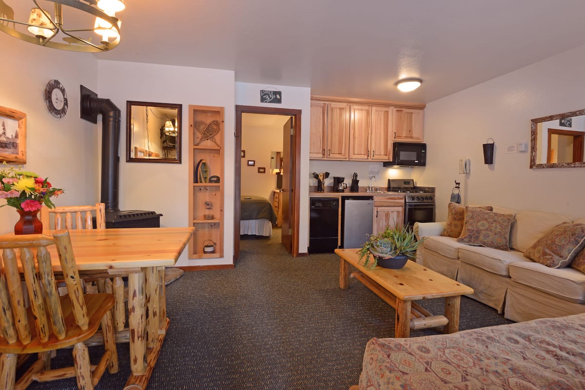 A cozy living room with a wooden dining table, a sofa, and a coffee table. There is a kitchen area with wooden cabinets and a microwave.