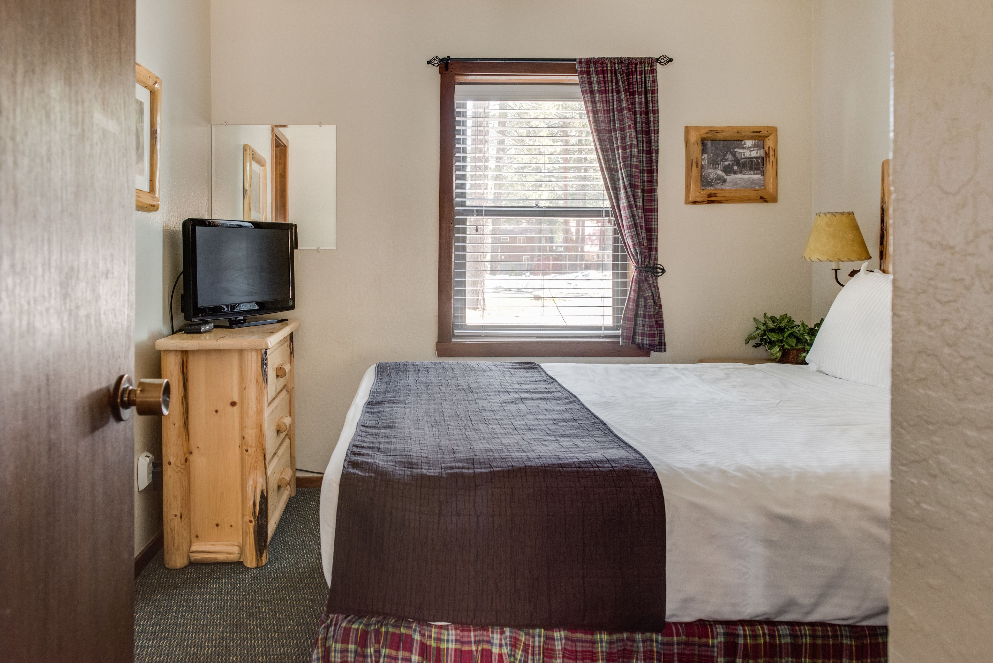Un dormitorio con cama, televisor sobre un tocador de madera, ventana con cortinas, lámpara y un cuadro en la pared.