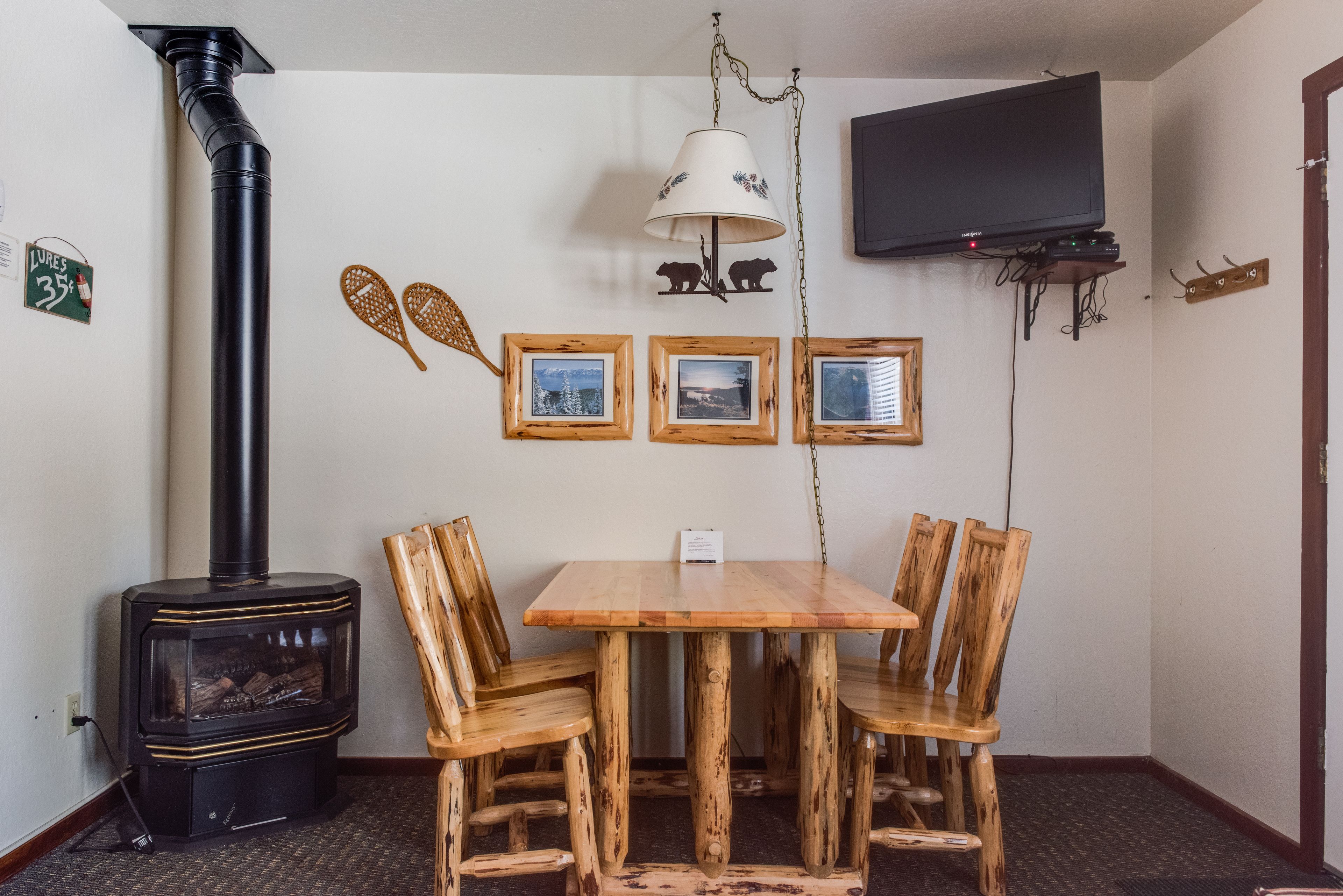 A room with a wood-burning stove, a wooden dining table with four chairs, a mounted television, and framed pictures on the wall.