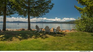 Vista sobre un lago con tres sillas Adirondack, cielo despejado y montañas a lo lejos.