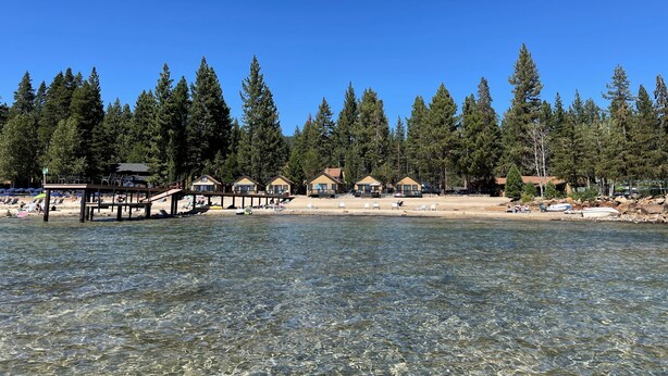 Una propiedad frente a un lago con aguas cristalinas, una playa de arena y varias cabañas rodeadas de altos pinos.