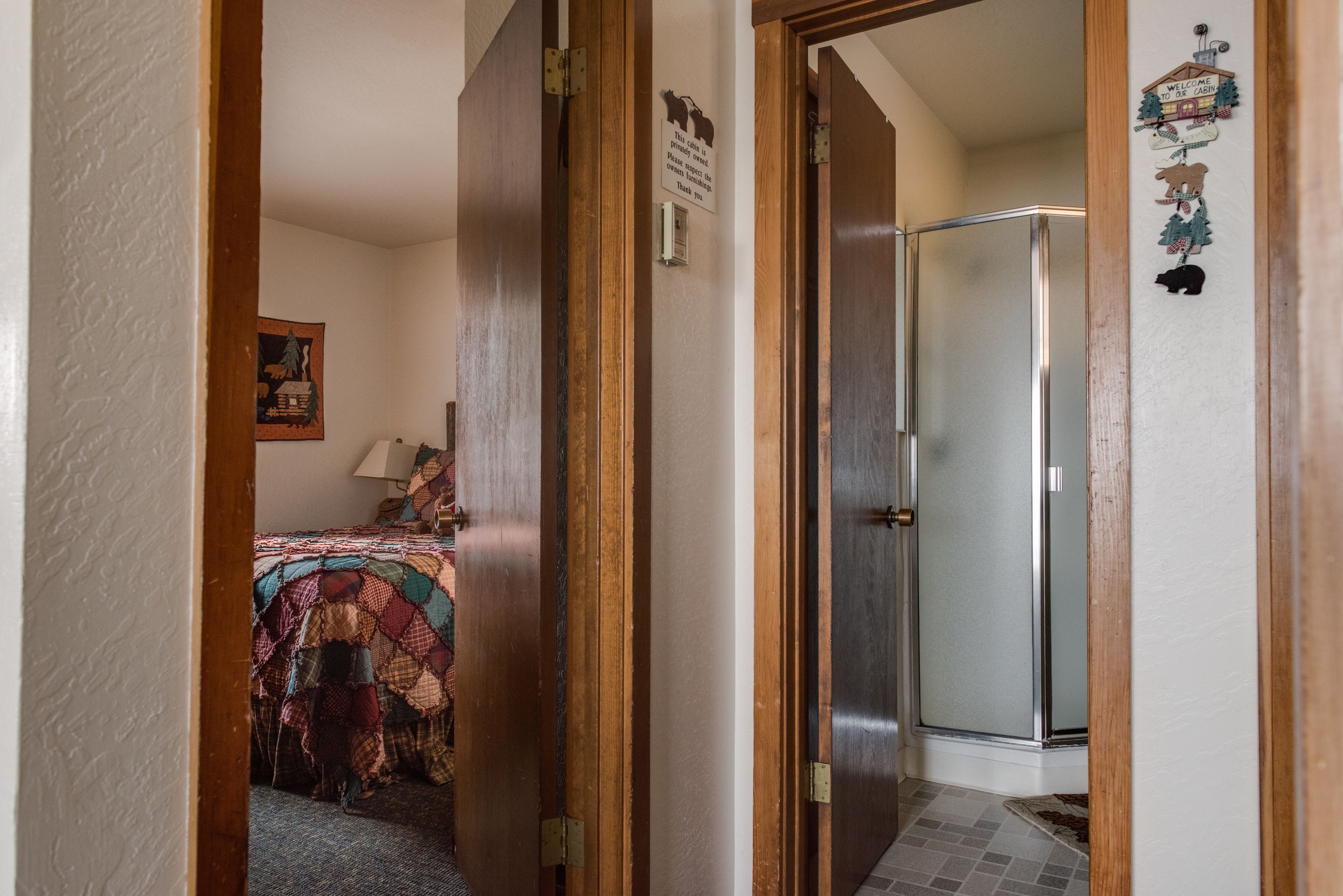 A narrow hallway with a wooden door leading to a bathroom with a glass shower. A bed with a colorful quilt is visible in the adjacent room.